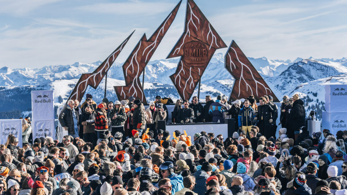Bei einem Musik-Open-Air-Event mit einer DJ-Kabine vor einem zerklüfteten, bergförmigen Schild versammelt sich eine große Menschenmenge.