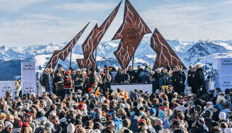 Bei einem Musik-Open-Air-Event mit einer DJ-Kabine vor einem zerklüfteten, bergförmigen Schild versammelt sich eine große Menschenmenge.