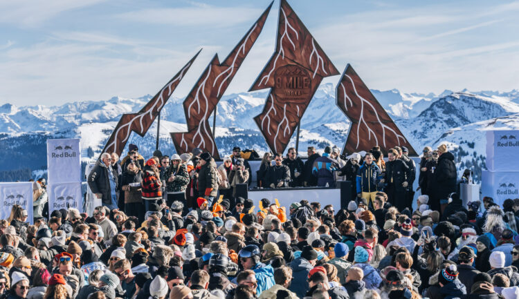 Bei einem Musik-Open-Air-Event mit einer DJ-Kabine vor einem zerklüfteten, bergförmigen Schild versammelt sich eine große Menschenmenge.