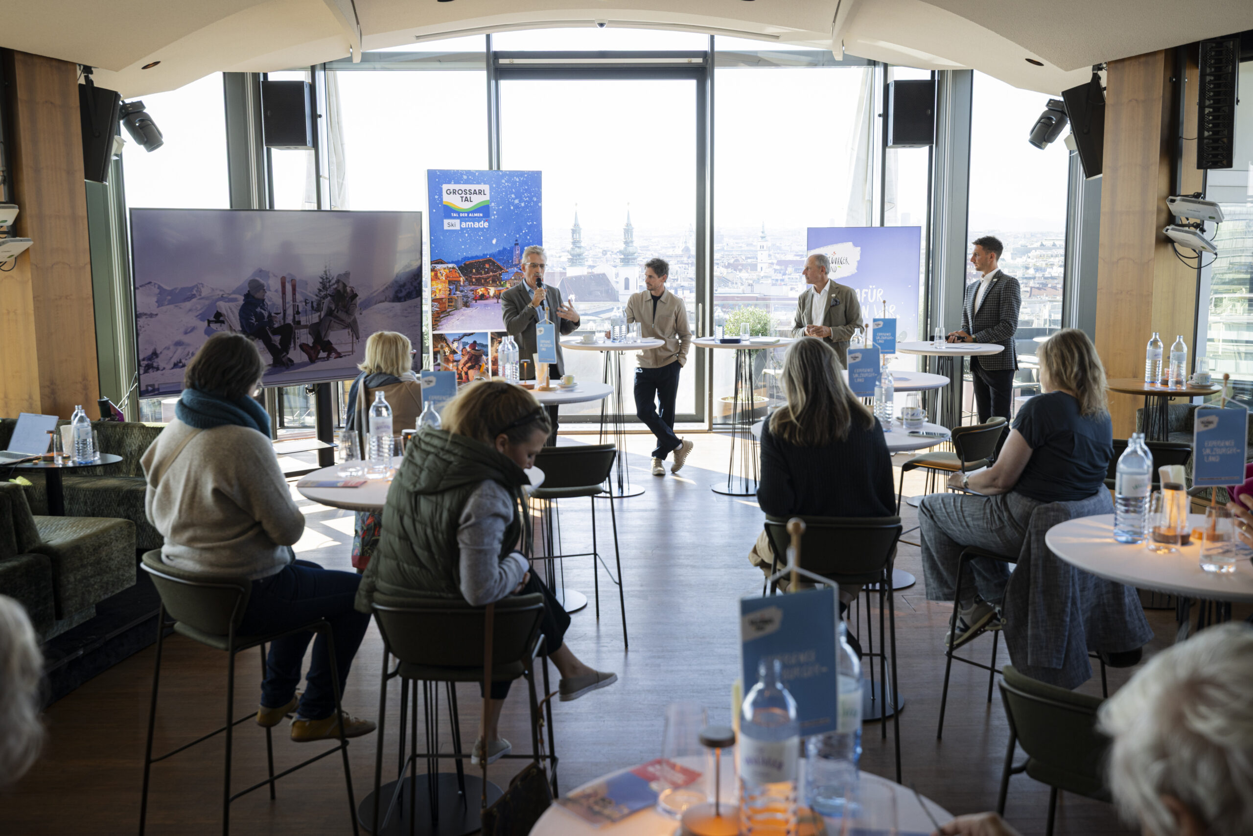 Vier Redner stehen bei einer Podiumsdiskussion in einem lichtdurchfluteten Raum, während das Publikum an hohen Tischen sitzt.